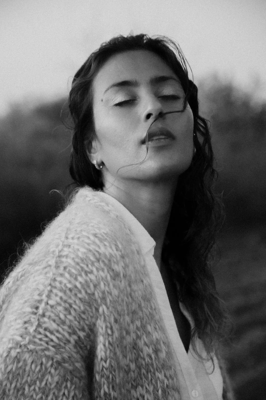 Black and white photo of a woman with long wavy hair, eyes closed, wearing a textured sweater, outdoors with a blurred background. 