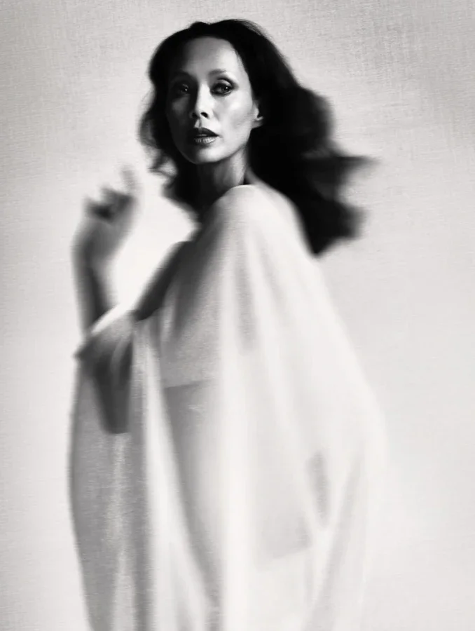 Black and white photo of a woman with flowing hair, wearing a light-colored garment, with a slightly blurred background.