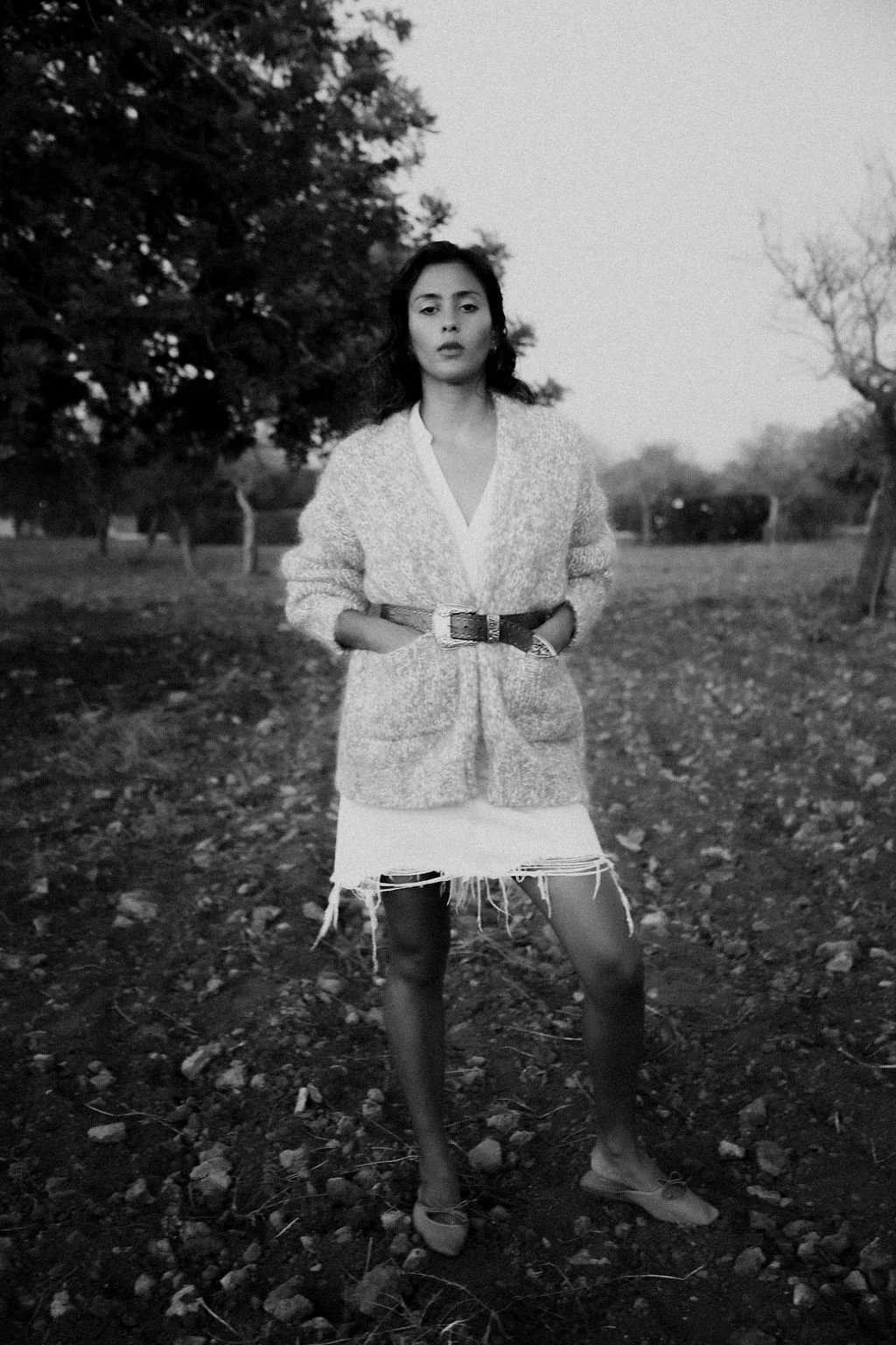 A young woman standing outdoors on textured ground with trees in the background, wearing a light-colored cardigan, a white dress, and flats, with her hands in her pockets, captured in black and white.