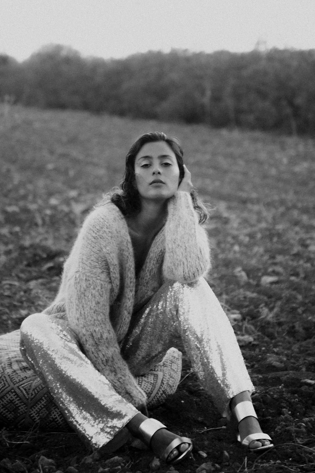 Black and white photo of a woman sitting outdoors on the ground with her head resting on her hand, wearing a cozy sweater and shiny, patterned pants.