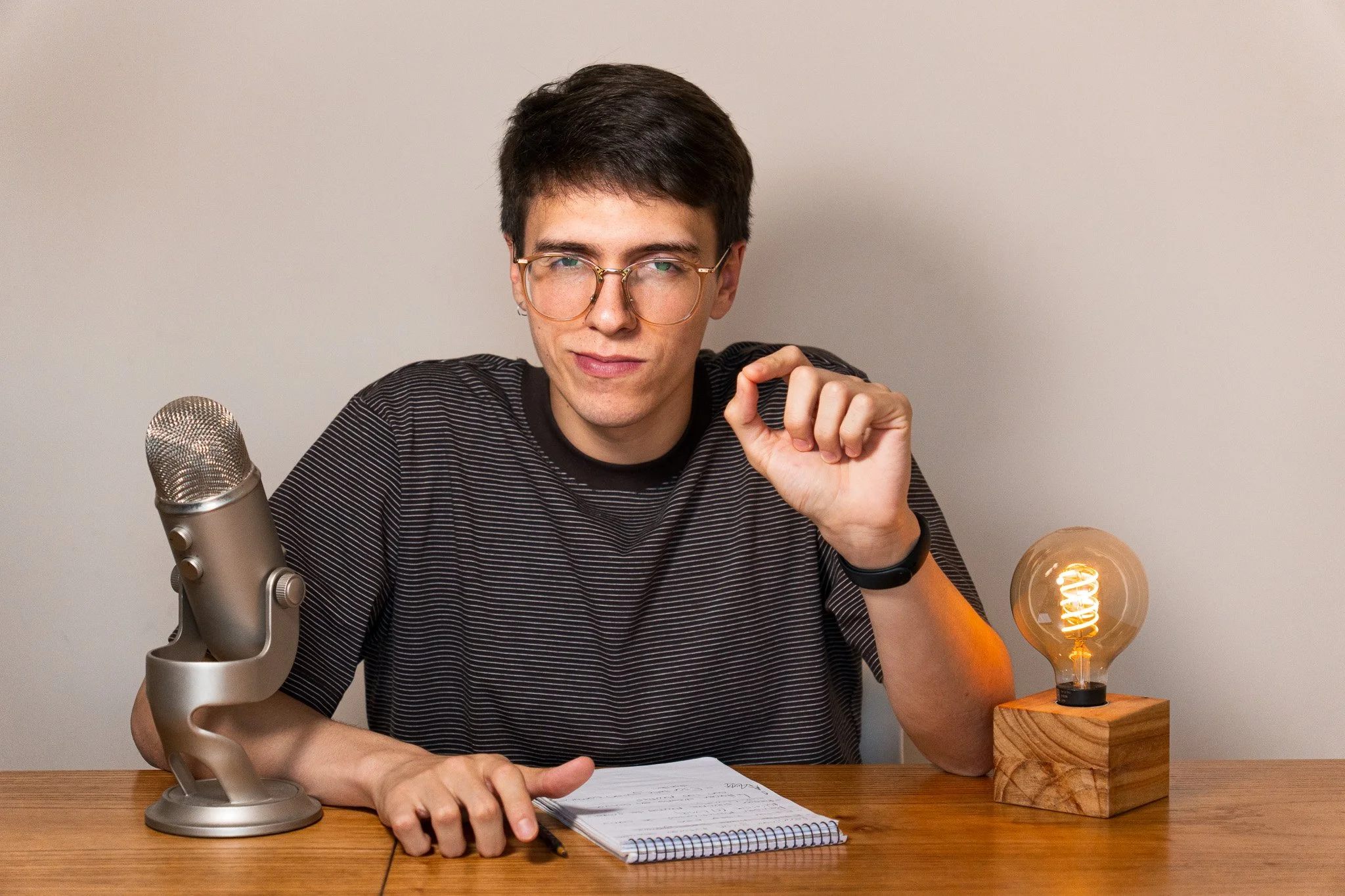 Joven hombre con gafas, sentado en una mesa de madera, sosteniendo un bolígrafo y una libreta, junto a un micrófono y una lámpara de luz incandescente, con fondo gris claro.