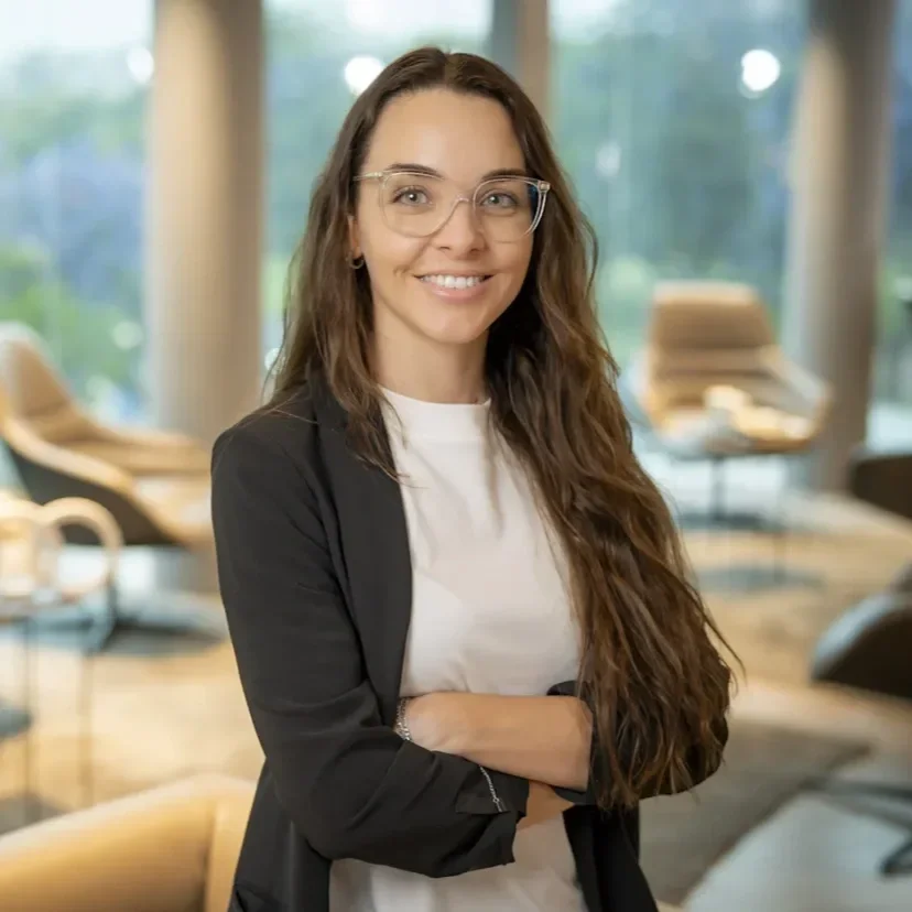 Mujer con gafas, cabello largo y rizado, sonriendo, en oficina moderna con sillones y libros en el fondo.