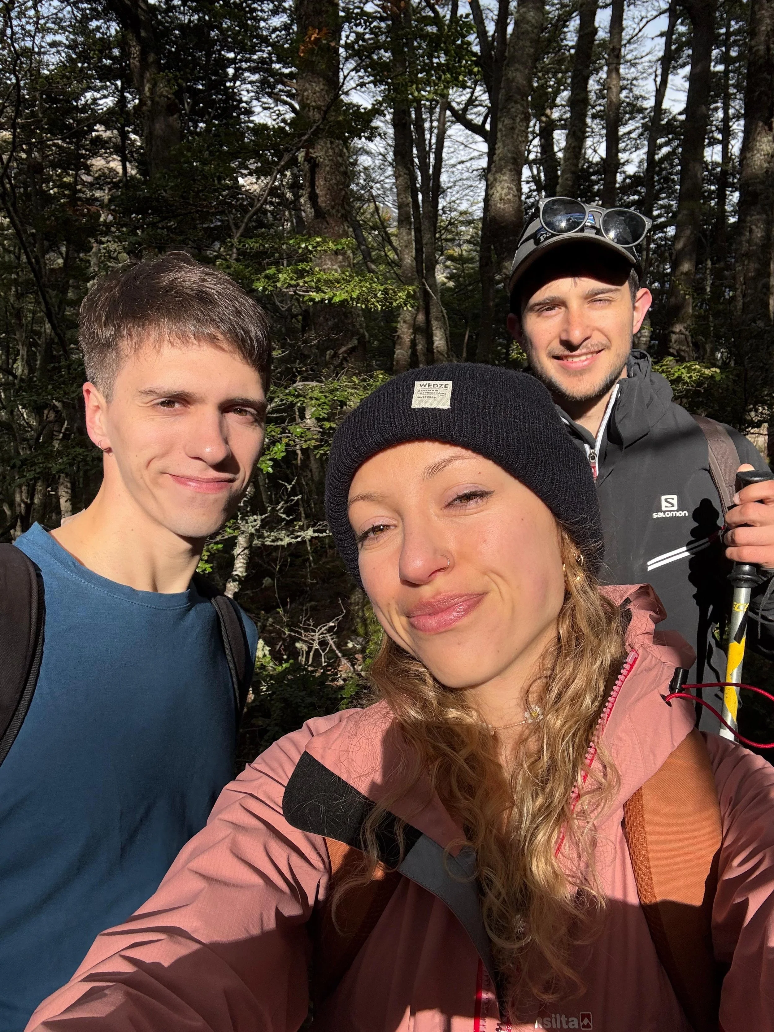 Tres personas en un bosque, dos hombres y una mujer, toman una selfie. Todos llevan ropa de abrigo y mochilas, parecen estar de excursión o caminando en la naturaleza.