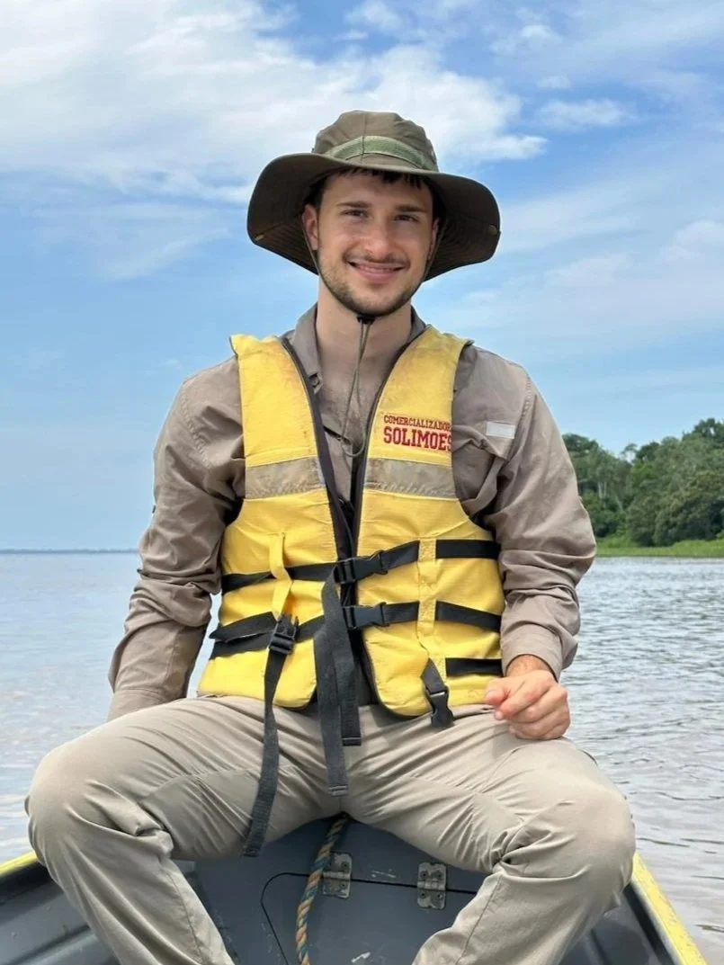 Hombre sentado en un bote en un río, usando sombrero de ala ancha y chaleco salvavidas amarillo.