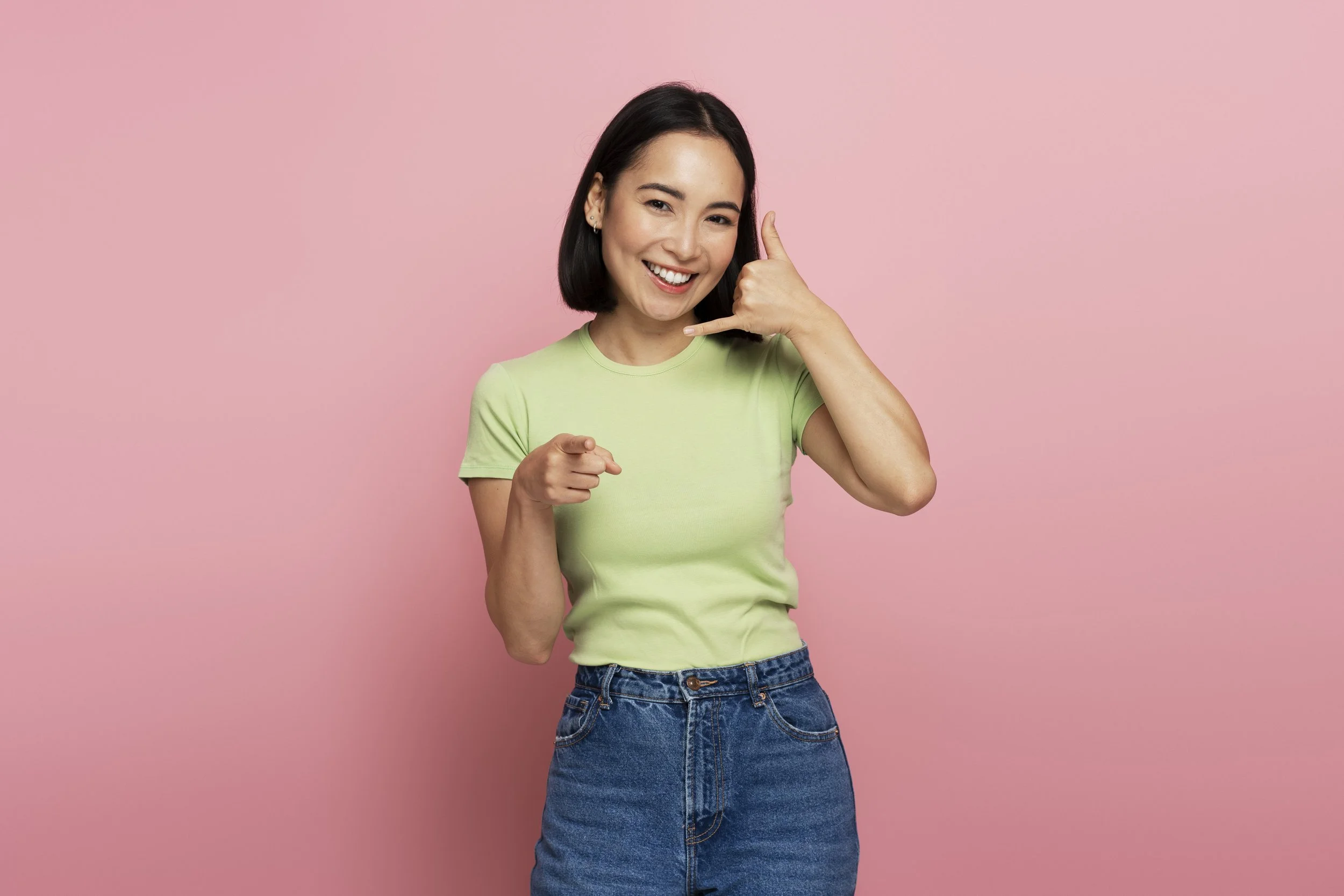 A young woman with shoulder-length dark hair smiling and making a phone call gesture with her hand while pointing at herself against a pink background.