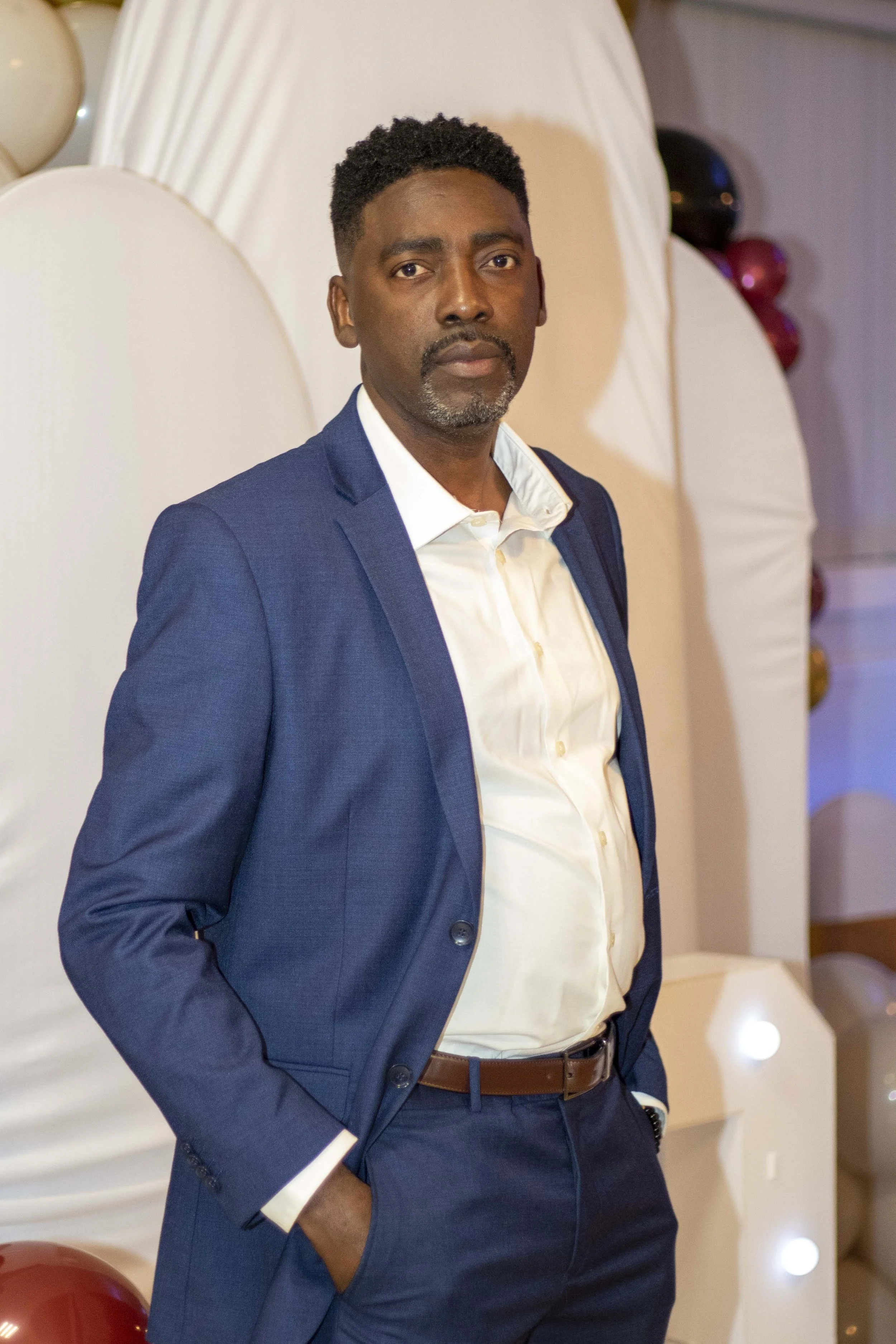 A man in a blue suit with white shirt and brown belt standing indoors in front of a decorated wall with balloons, looking at the camera with a neutral expression.