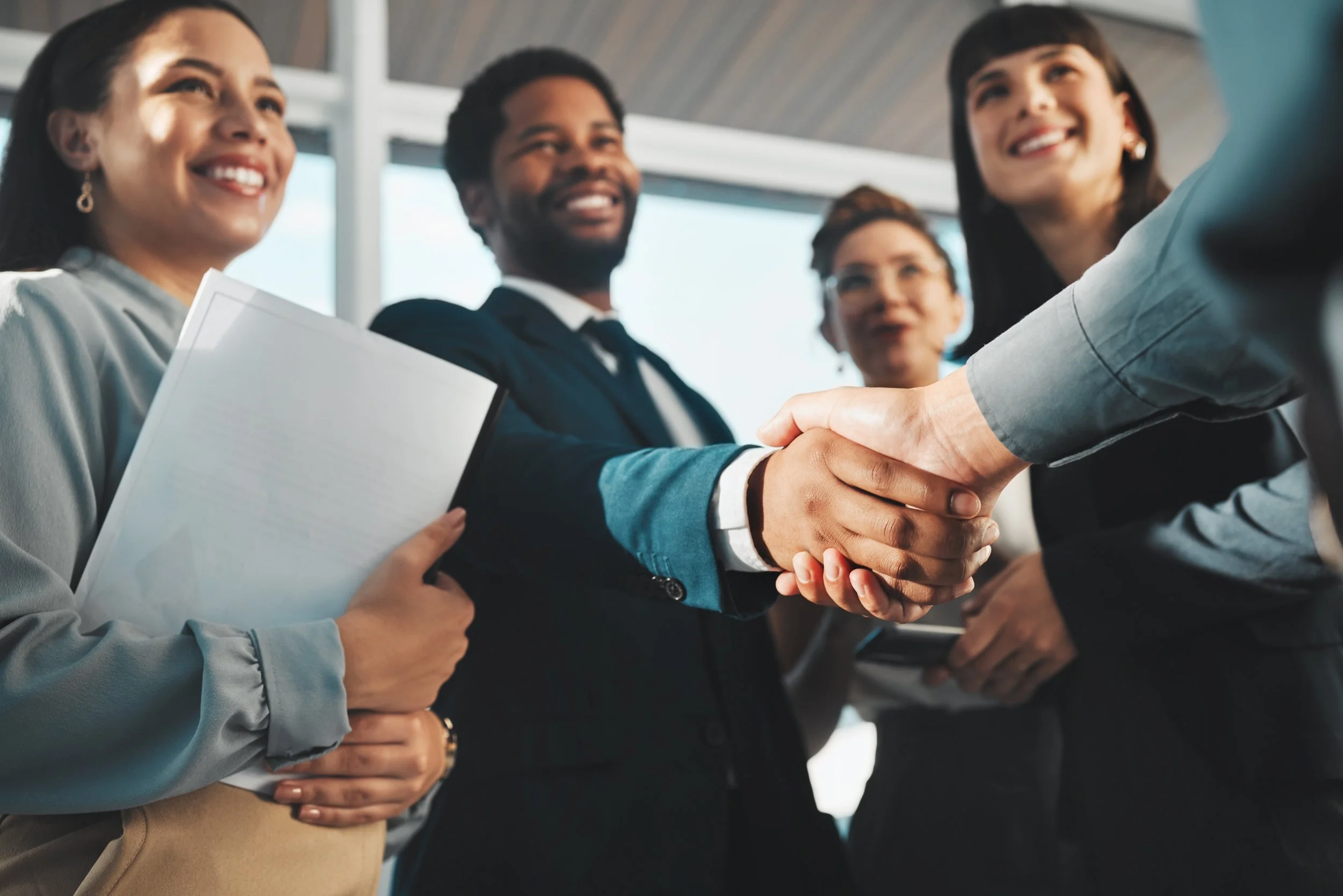 A group of diverse professionals inside an office or conference area, engaged in handshaking and smiling, indicating a positive and collaborative interaction.