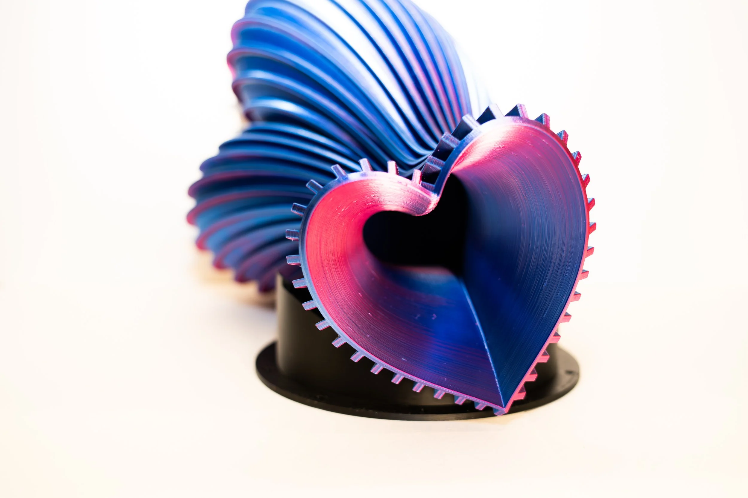 Metal gears shaped like a heart with vibrant pink and purple colors, mounted on a black stand