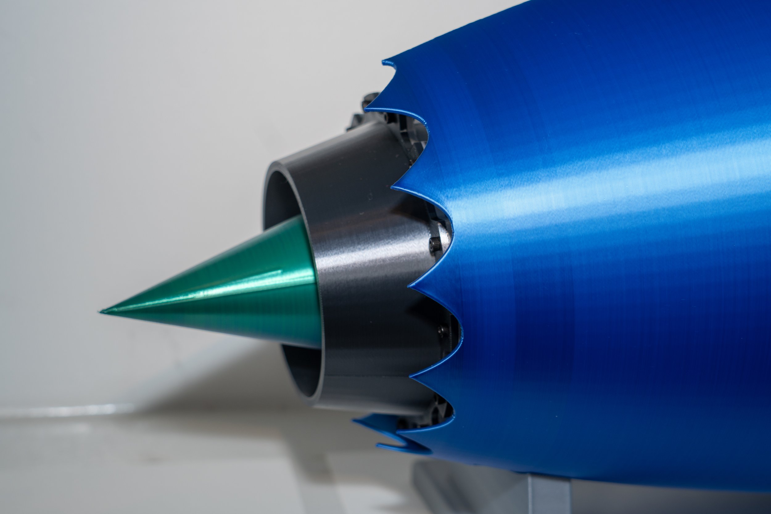 Close-up of a turbine engine or jet engine component with a green nose cone, metallic parts, and blue exterior shell with jagged edges.