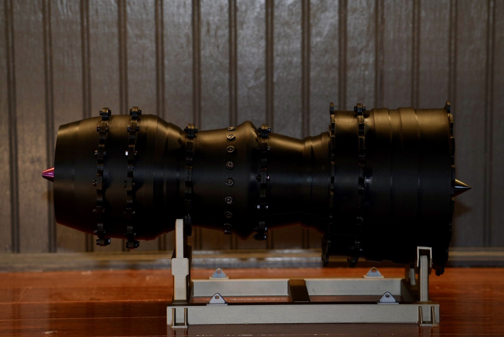 A large black spherical object with multiple segments and attached fasteners, mounted on a metallic stand, set against a wooden and panel background.