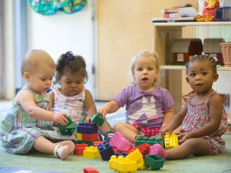 Four young children sitting on a floor playing with colorful plastic building blocks in a room that appears to be a play area.