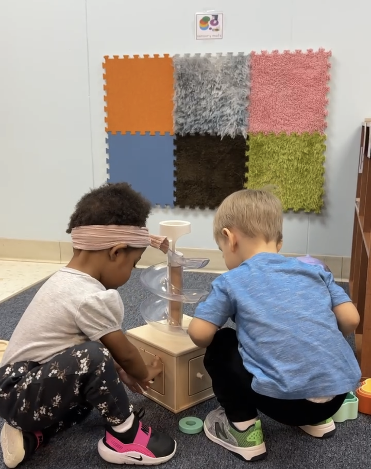 Two young children playing with a spiral marble run in a classroom, with colorful textured wall squares in the background.