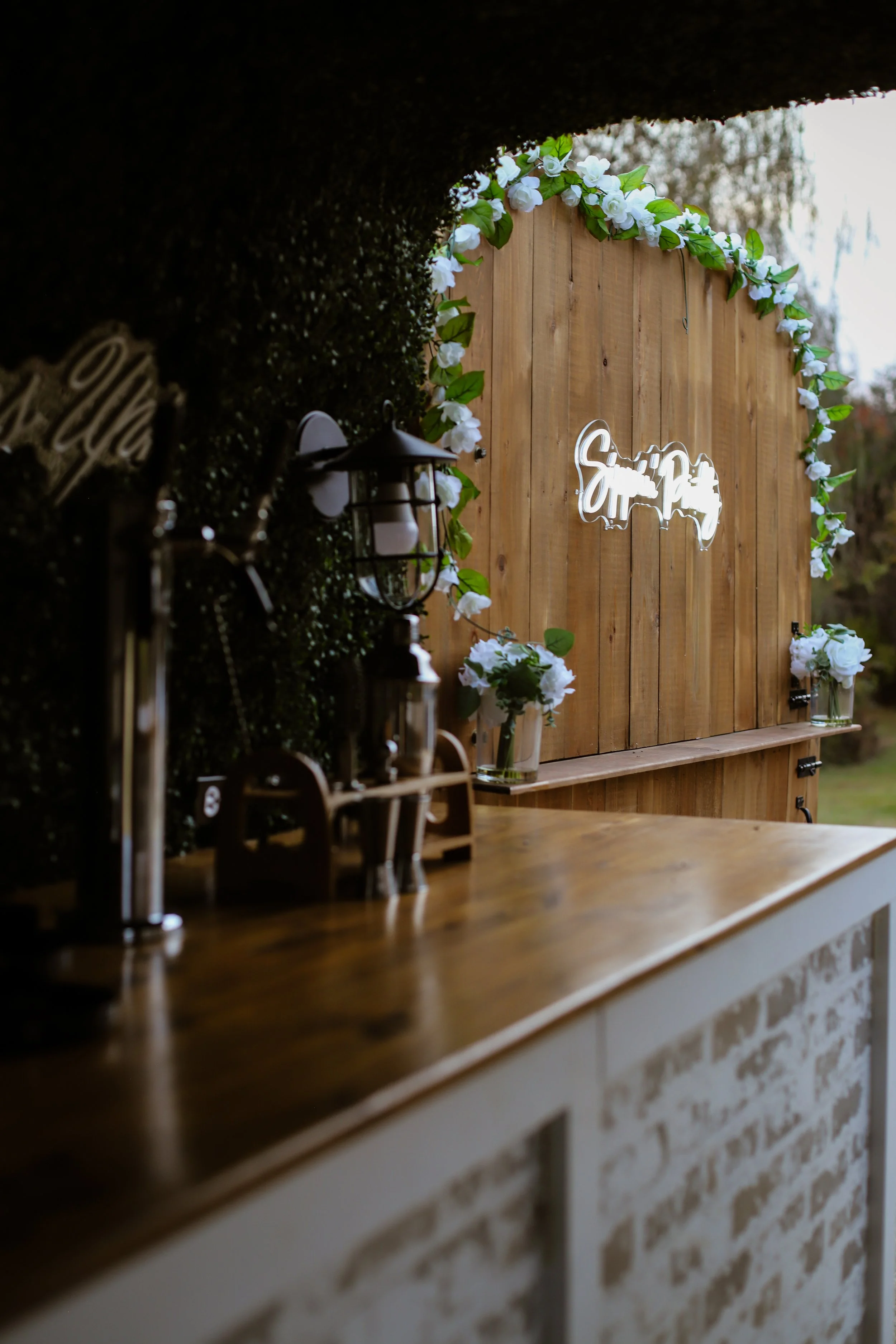 Outdoor wedding or event decor featuring a wooden backdrop decorated with white flowers and green leaves, and a neon sign with the word 'Smile'.