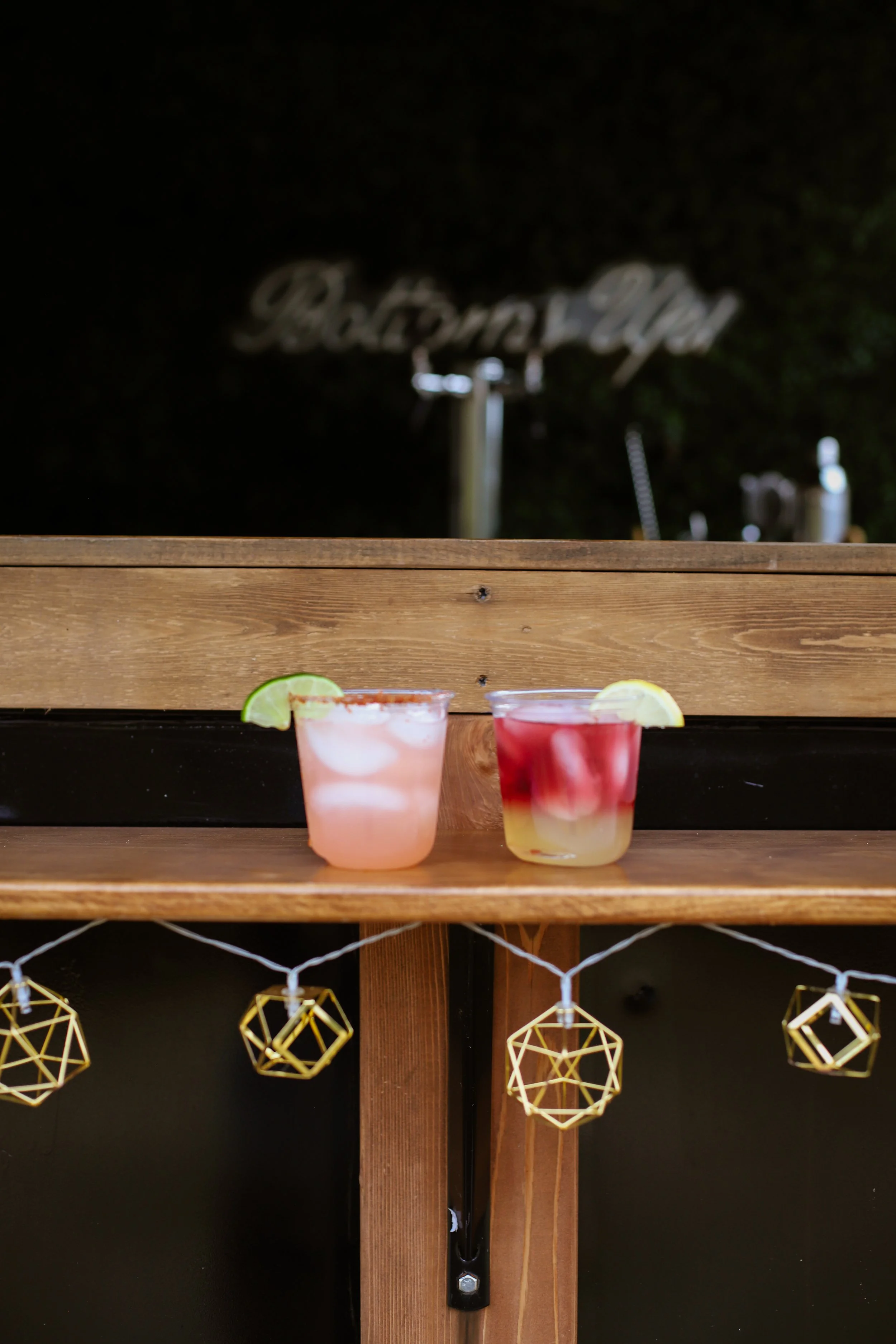 Two cocktails with lemon and lime slices on a wooden shelf, with blurry background.