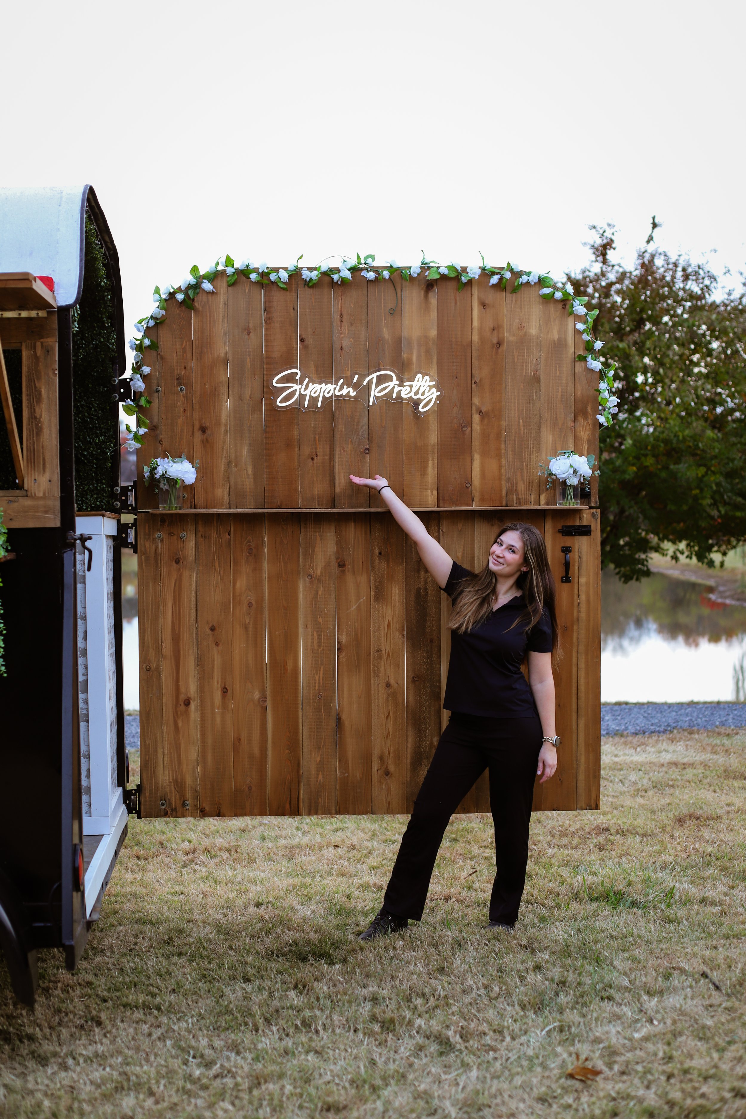 A woman standing outdoors near a lake, pointing at a wooden sign that says 'Gippin' Pretty' with decoration like flowers and greenery.