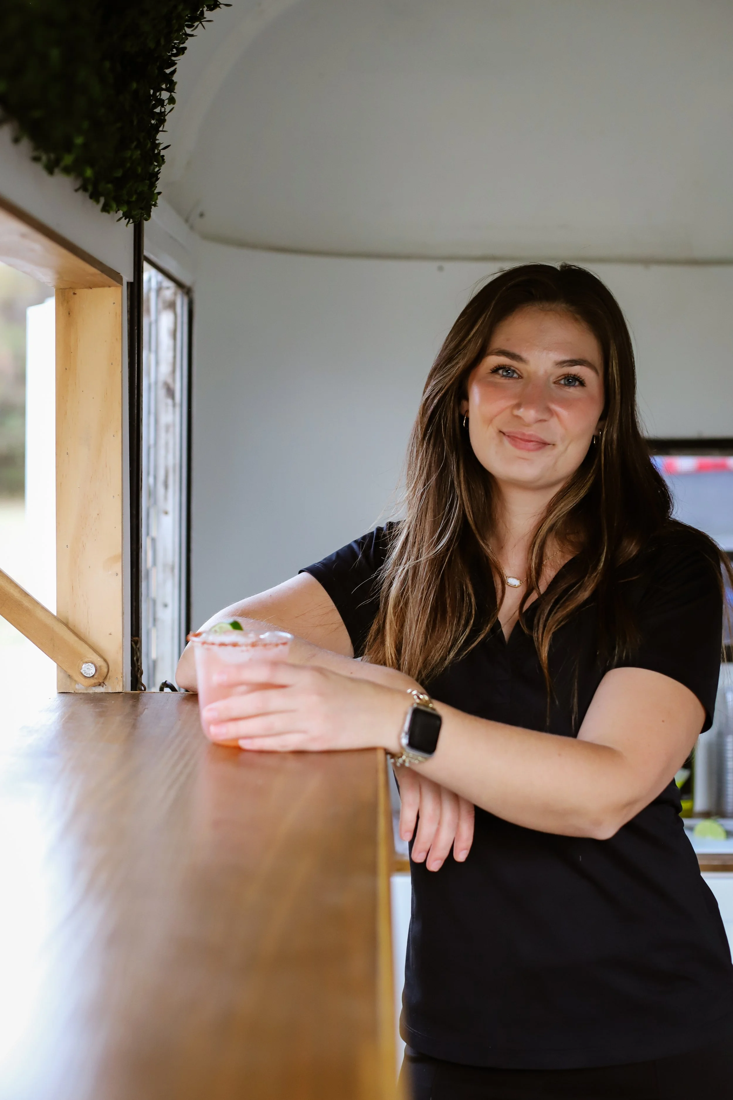 A young woman with long brown hair, wearing a black t-shirt and a smartwatch, smiling and leaning on a wooden counter with a pink cocktail in hand, inside a bright space with a window and greenery