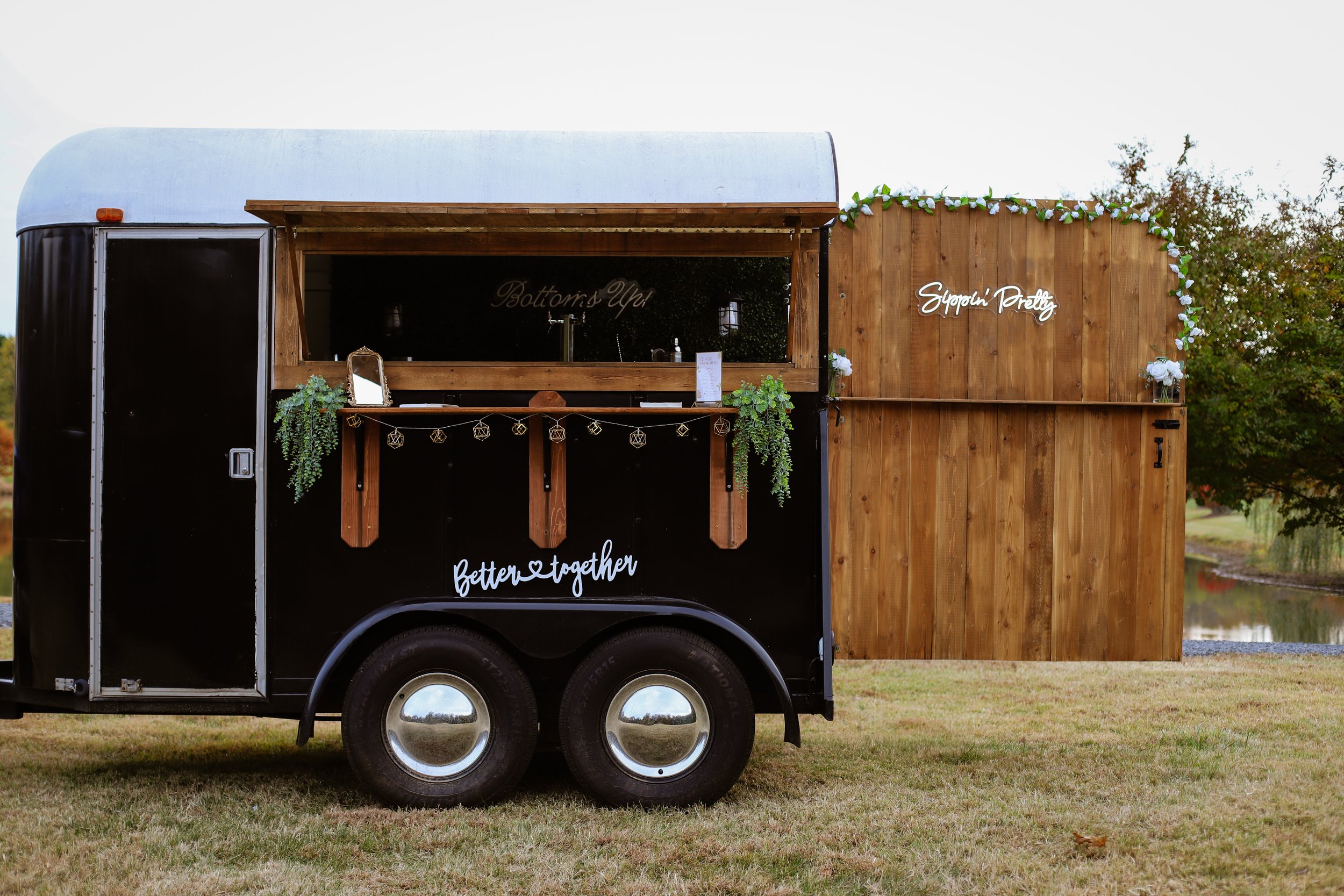 Black food truck with wooden accents and decorative plants, parked on a grassy area near a pond, with signs reading 'Better together' and 'Supportin' Pretty'.