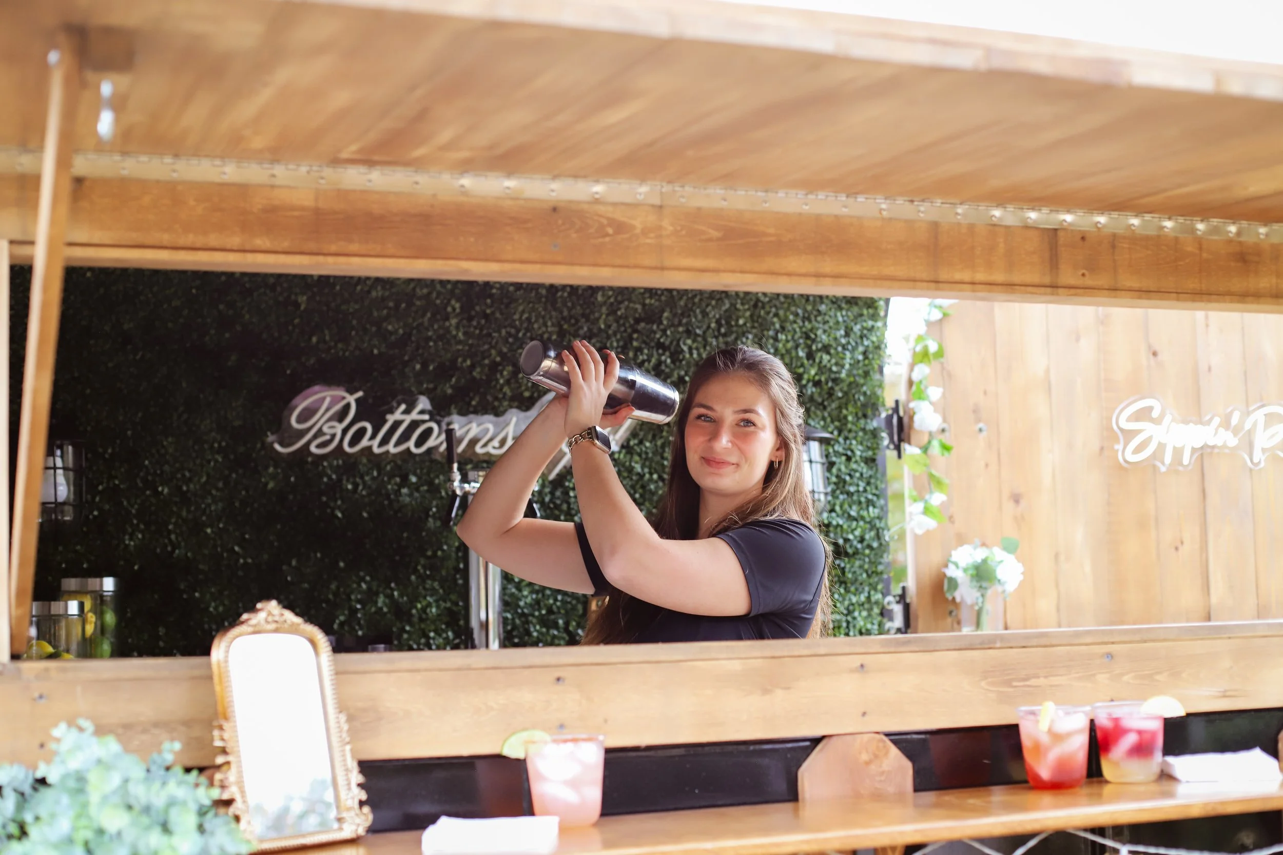 A young woman smiling as she prepares a cocktail behind a bar with a mirror in a wooden frame, flanked by greenery and a sign that says 'Bottoms'. She is holding a cocktail shaker, wearing a black shirt, and has long brown hair.