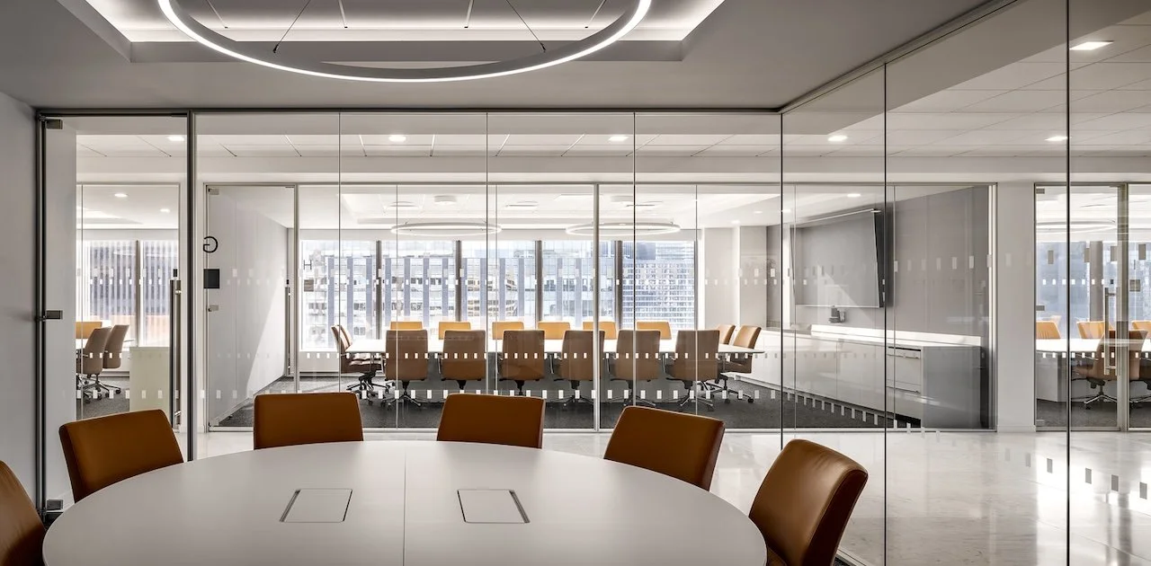 Large round meeting table in a glass room overlooking the boardroom with a large V-shape wood veneer conference table.