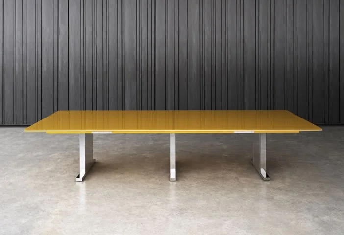 A modern, rectangular yellow table with metal legs, set against a black wood-paneled wall and a concrete floor.