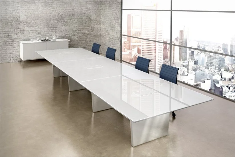 Modern office conference room with a long white table, four blue chairs, a brick wall, large windows with city views, and a white sideboard