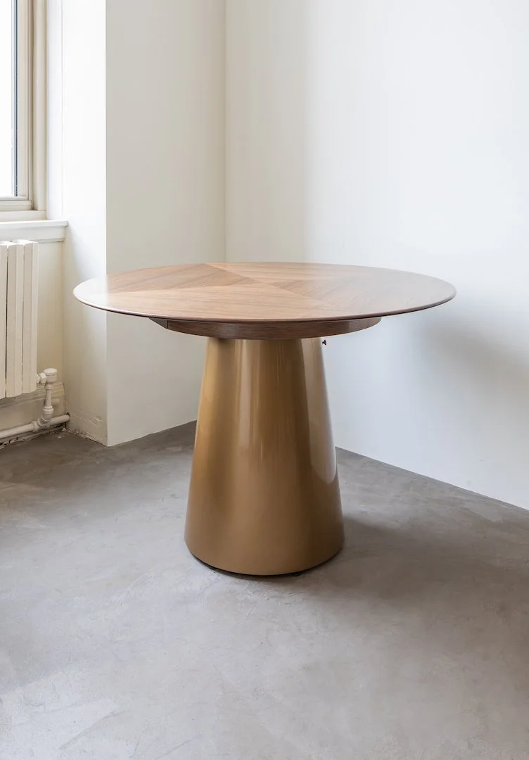 Round wooden table with a gold cone-shaped base in a minimalistic room with concrete floor and white walls.