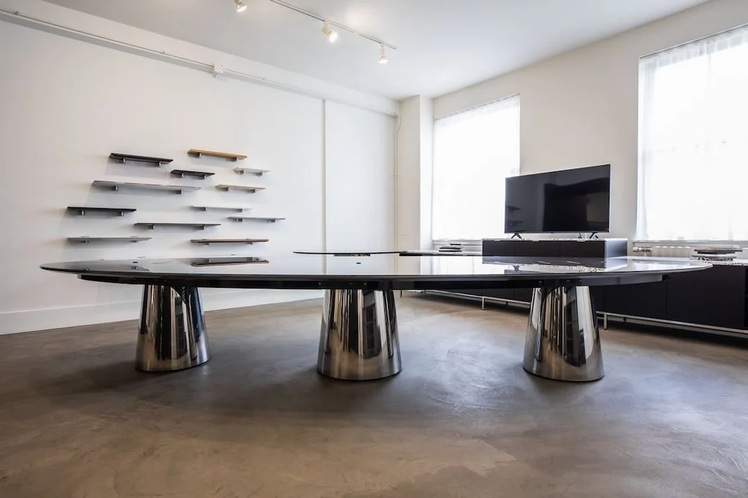 Modern, minimalist office or conference room with a large black oval table with metallic bases, a flat-screen TV on a black stand, wall-mounted shelves arranged in a pattern, and large windows with white curtains allowing natural light.