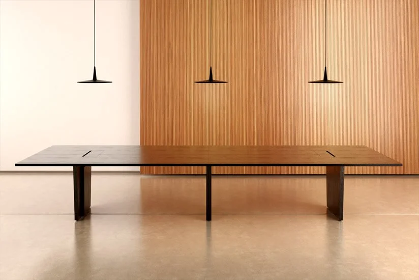 Minimalist interior with a long wooden conference table, three black pendant lights hanging above, and a wooden-paneled wall in the background.