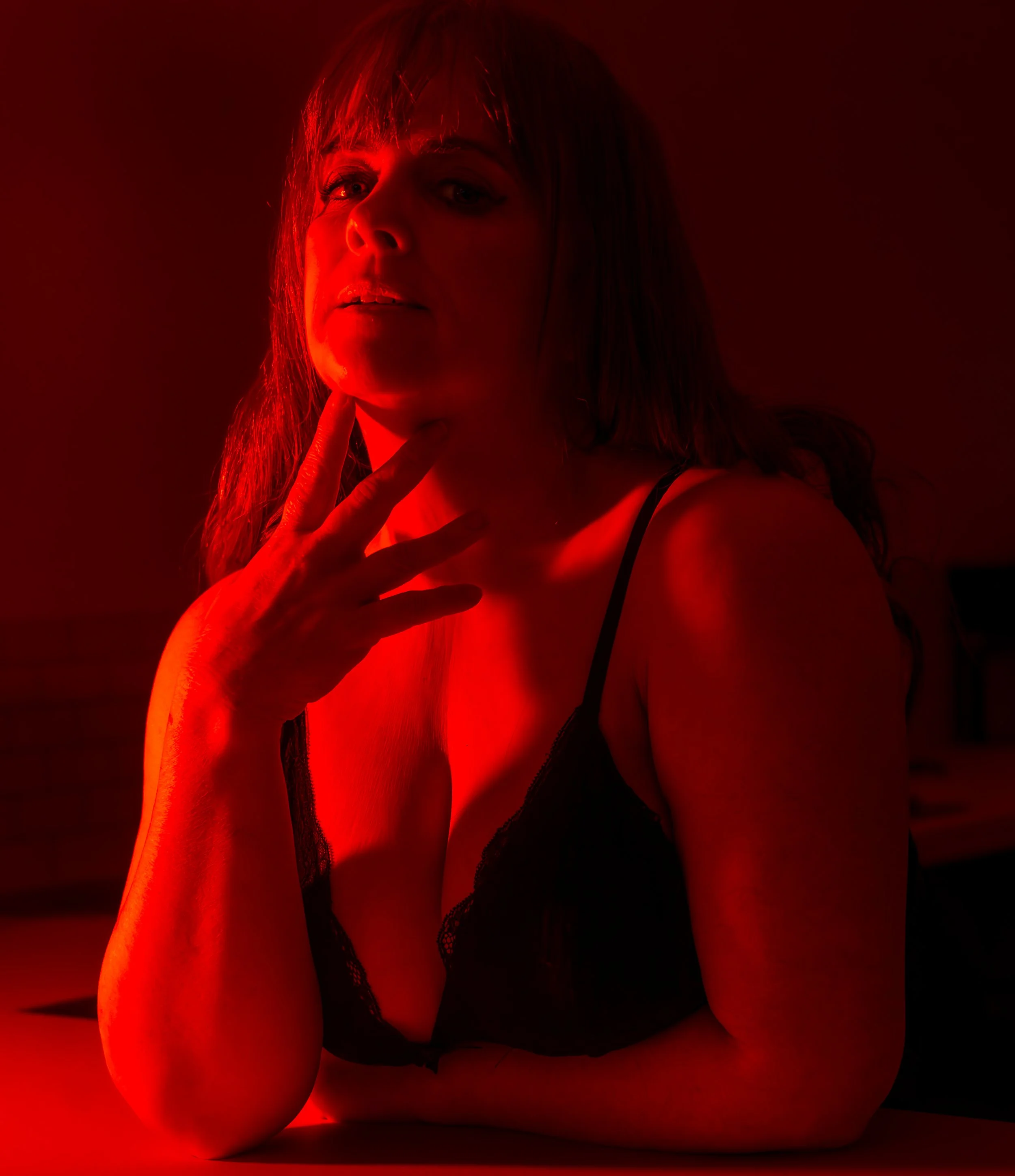 Relaxed boudoir portrait in home setting
A woman with long hair wearing a black lace top, illuminated by red lighting, sitting with her hand near her face.