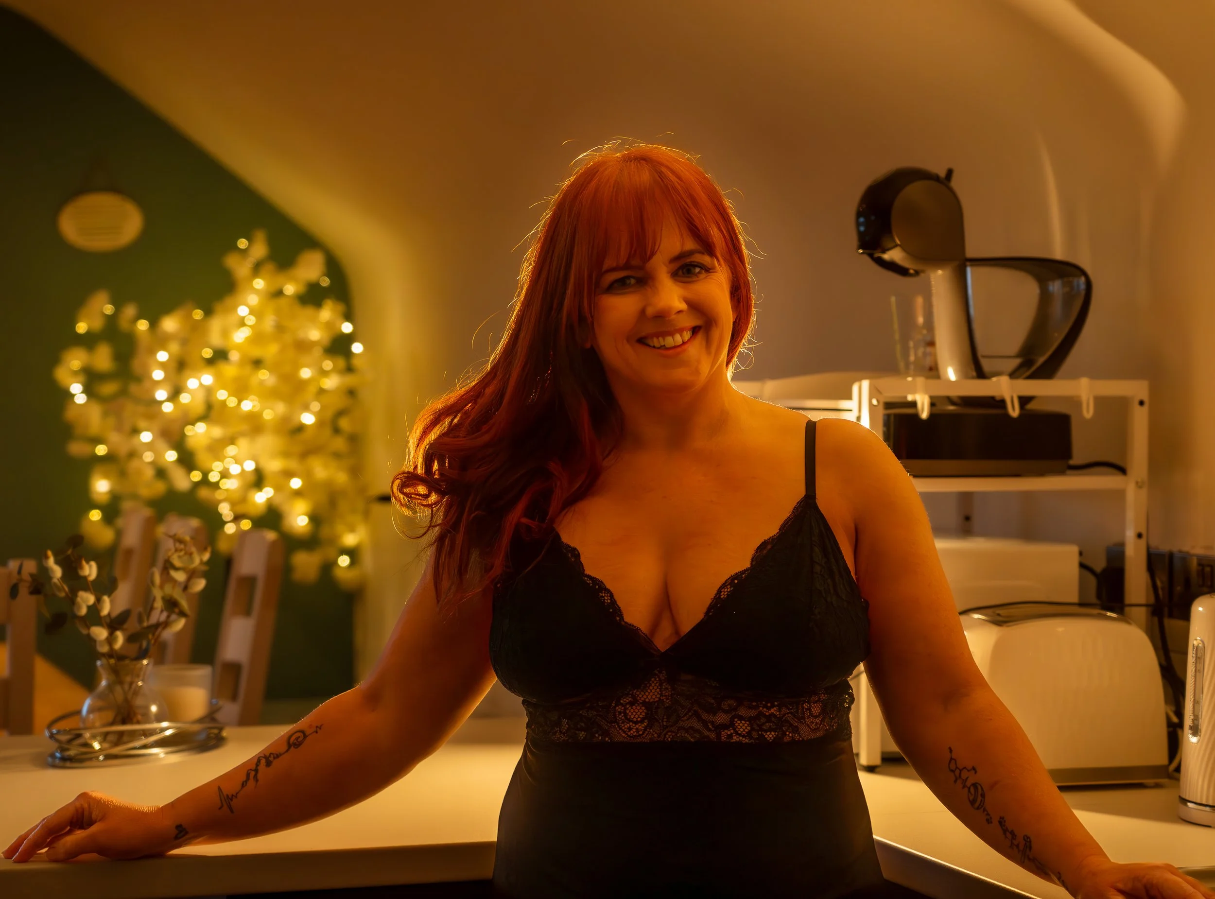 Natural light boudoir photography Northern Ireland, A smiling woman with red hair wearing a black lace top stands in a warmly lit room, with a decorated tree in the background.
