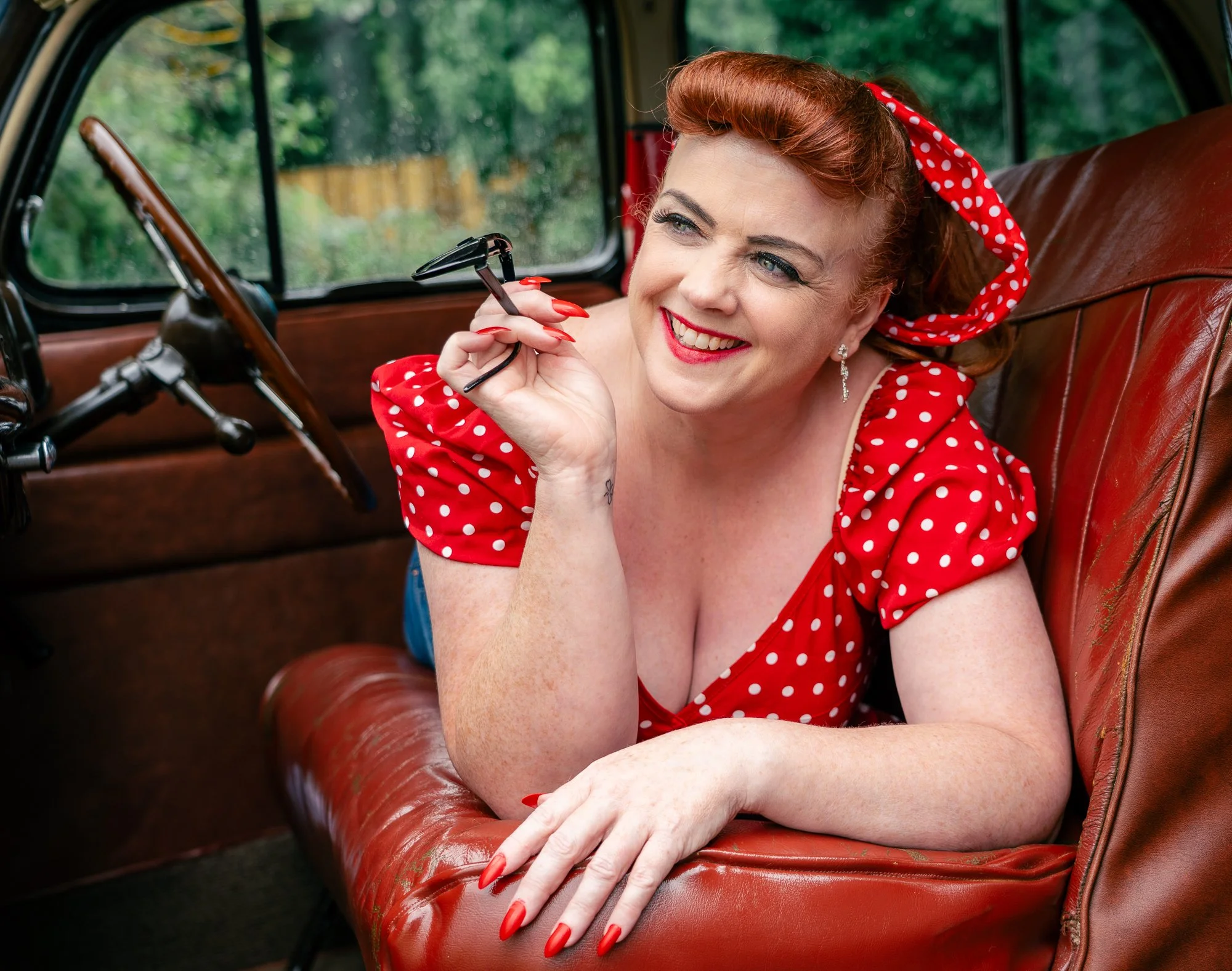 Relaxed 50's pin up boudoir portrait in home setting
A woman with red hair, polka-dot dress, and matching headscarf leaning on a vintage car's leather seat, smiling and holding glasses.