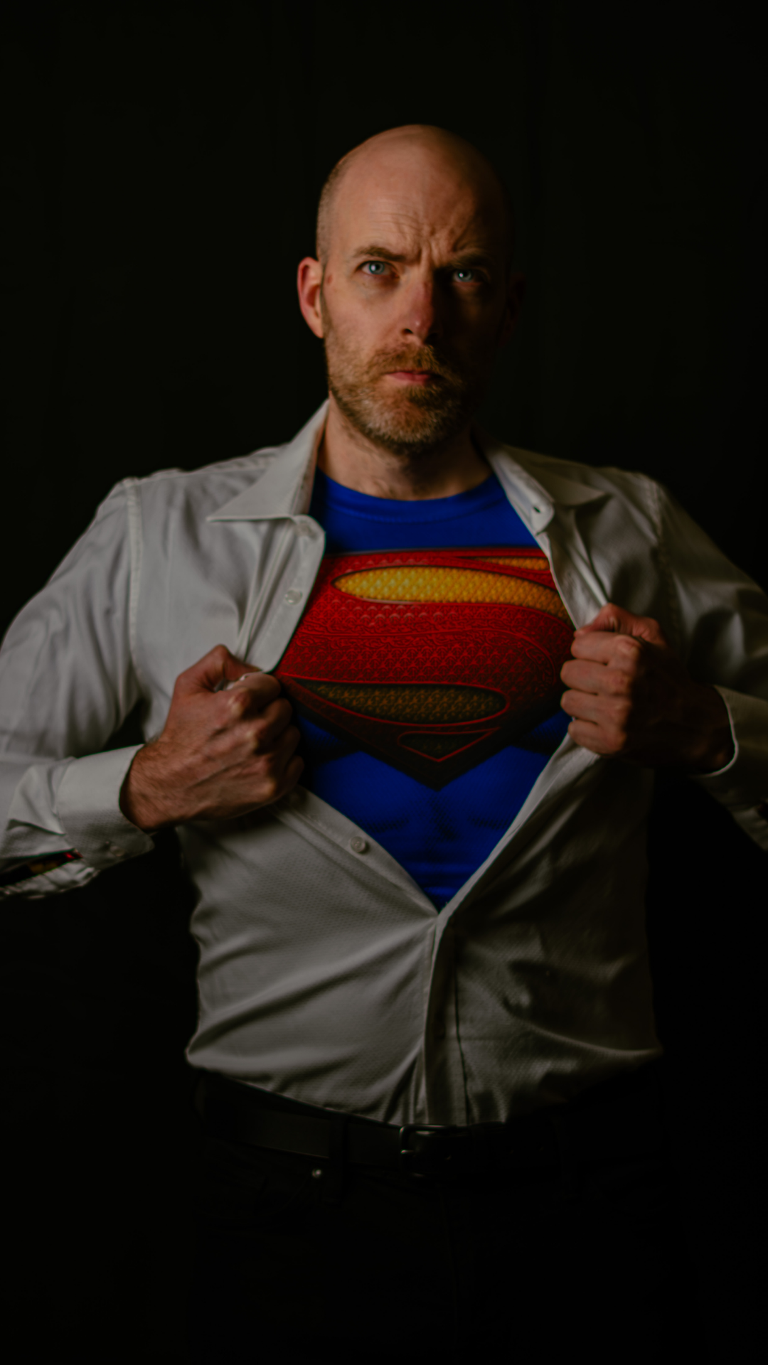 A man with a bald head and beard opening his white dress shirt to reveal a Superman logo t-shirt underneath. He has a serious expression and is set against a dark background. omagh Liam o Neill