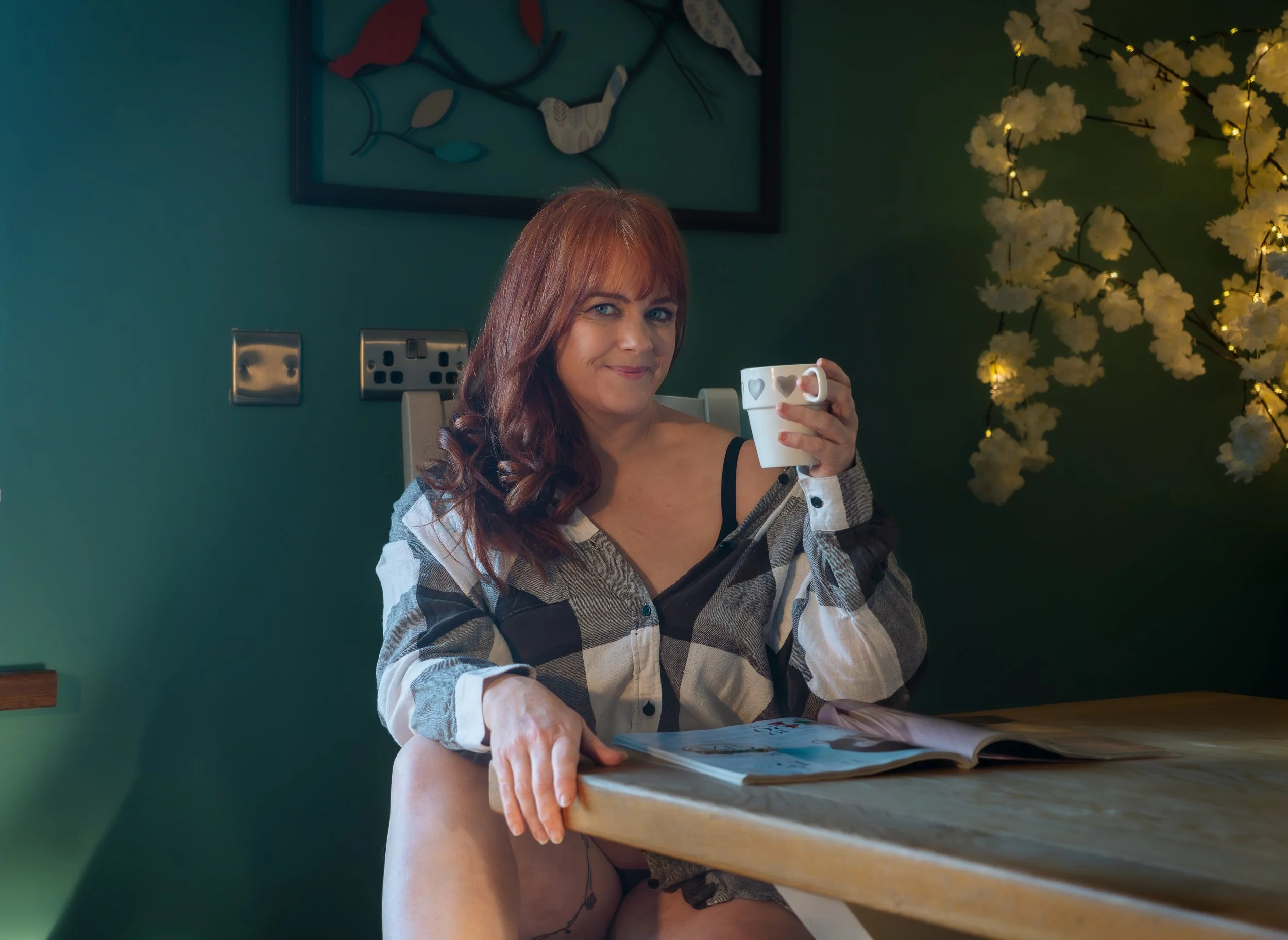 Natural light boudoir photography Northern Ireland, a woman with red hair sitting at a wooden table, holding a white mug with heart designs, smiling at the camera