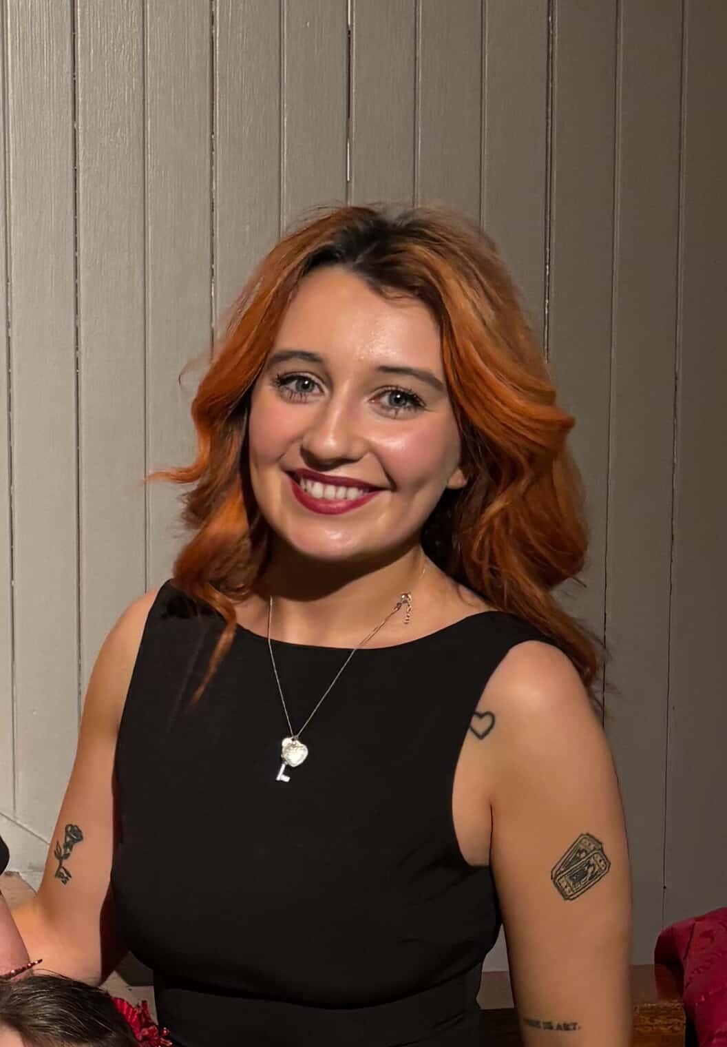 A smiling woman with shoulder-length red hair wearing a sleeveless black top and a heart-shaped necklace standing in front of a wooden wall. boudoir confidence experience northern ireland