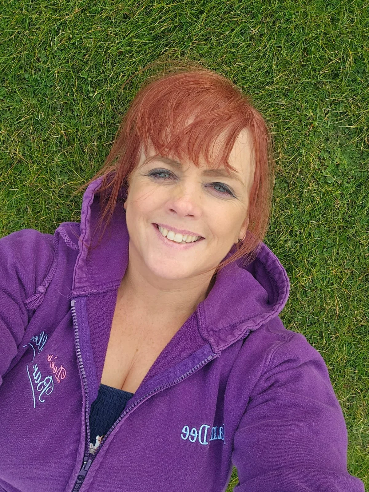 A woman with red hair lying on green grass, smiling, wearing a Dee's Barbelles purple hoodie. boudoir transformation omagh