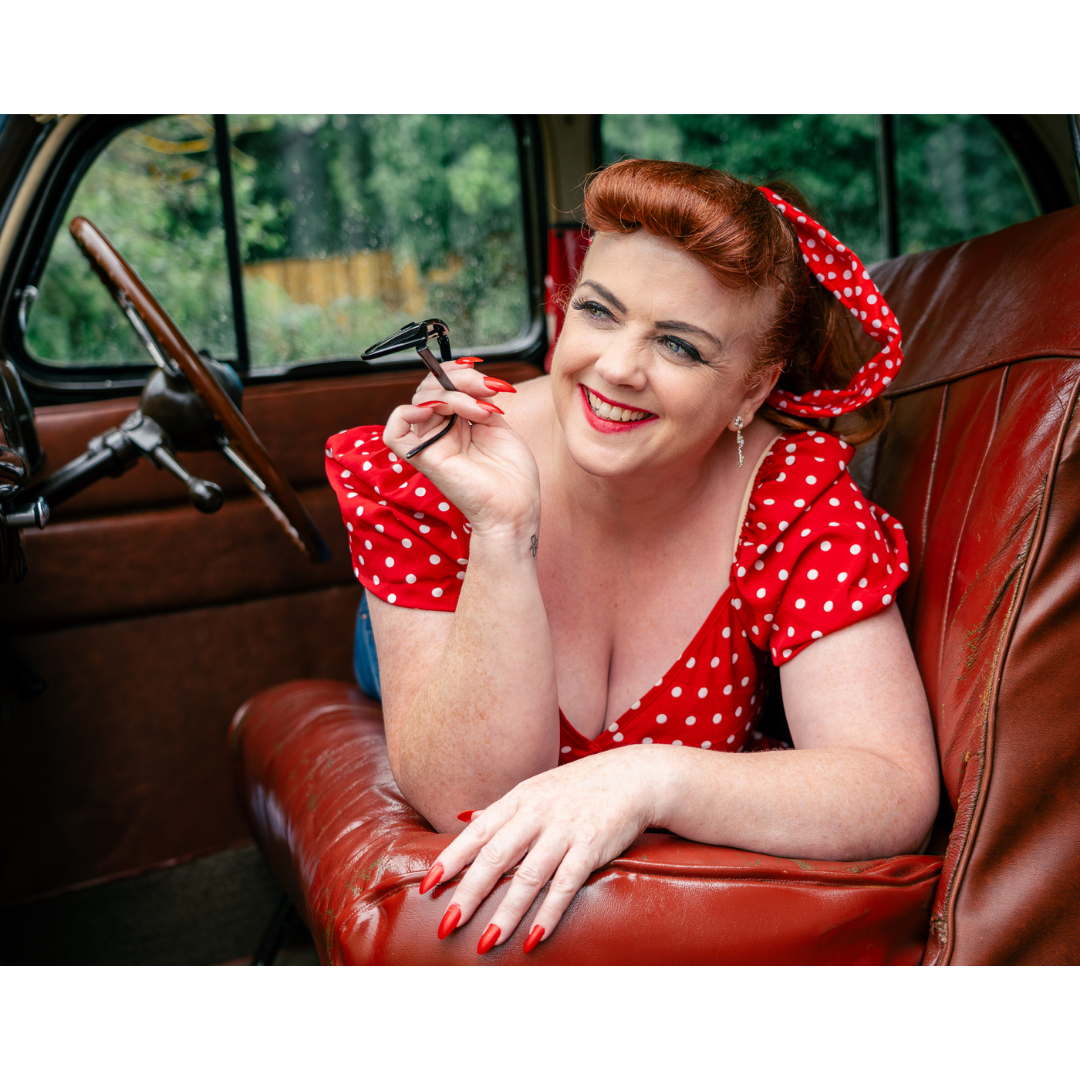 A woman with red hair, wearing a red polka dot dress and matching hair bow, is sitting inside a vintage car, smiling, holding a glasses, with her arm resting on the door. boudoir transformation omagh
