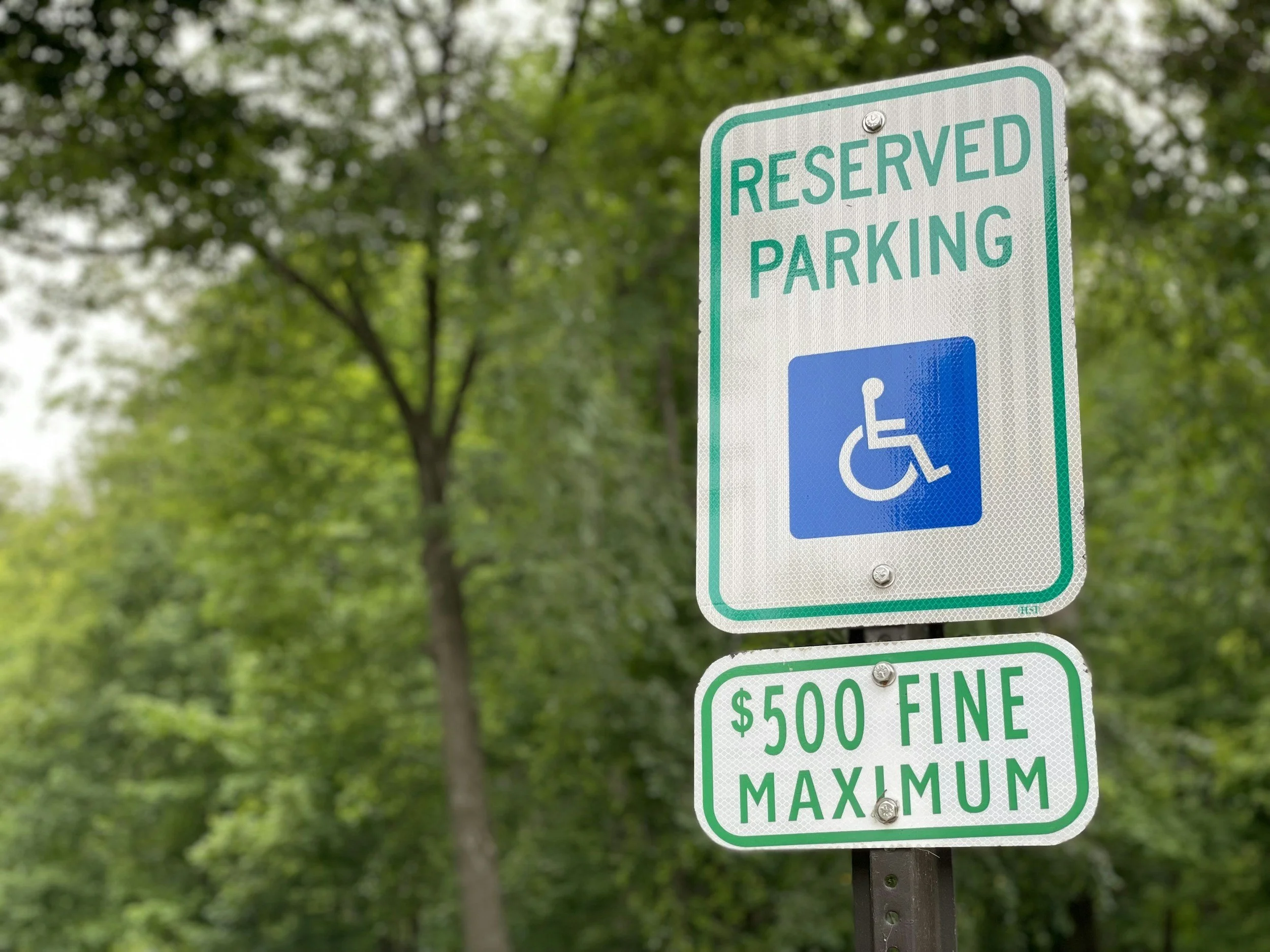 A sign indicating reserved parking for disabled with a $500 fine maximum, set against a background of green trees.
