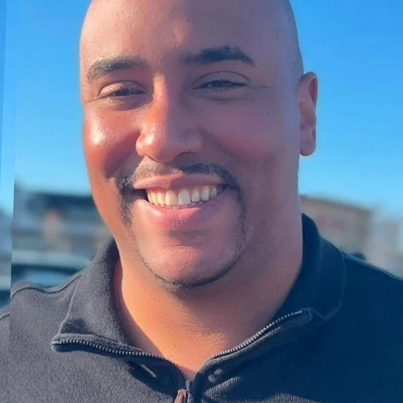 A smiling man with a mustache and goatee, wearing a black zip-up jacket, standing outdoors on a sunny day.