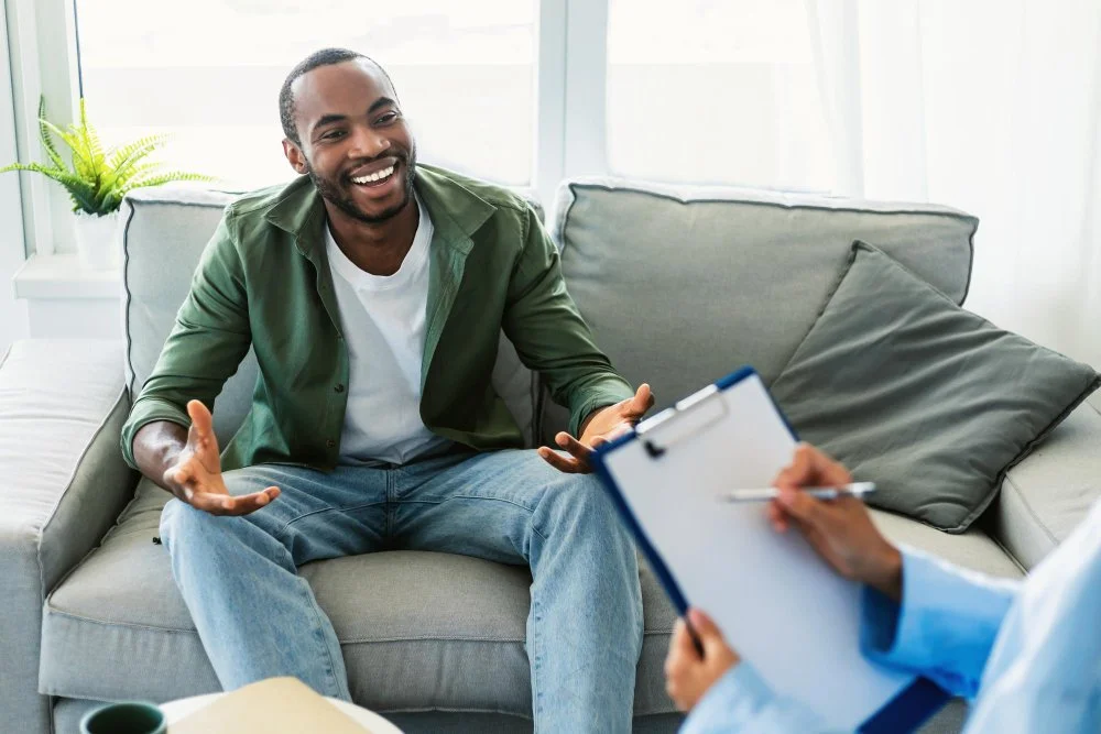 A man smiling and engaging in conversation with a therapist or counselor, who is taking notes on a clipboard.