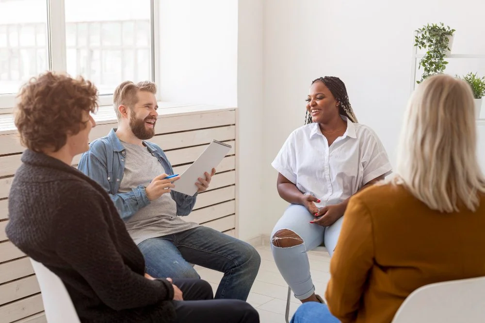 Four diverse people sitting in a circle and smiling in a therapy or support group session in a bright room with large windows and green plants.