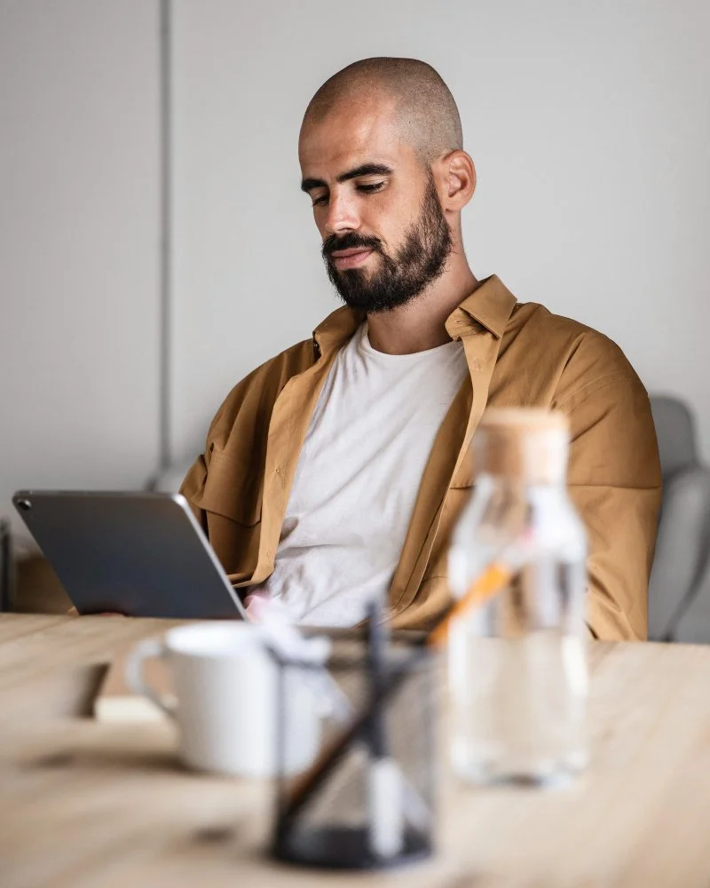 Man with a beard and shaved head sitting at a table, looking at a tablet in his hands, with coffee, water, and office supplies on the table, in a modern office setting.