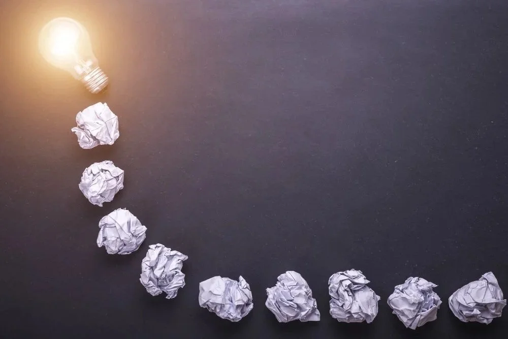 Line of crumpled papers forming a rising curve leading to a lit light bulb.
