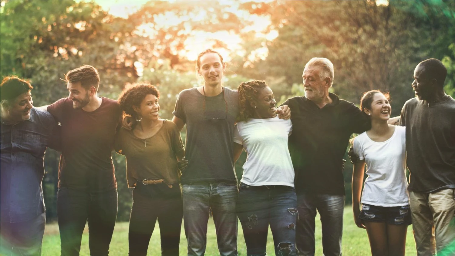 Group of diverse people standing together outdoors in a park, smiling and linking arms during sunset.