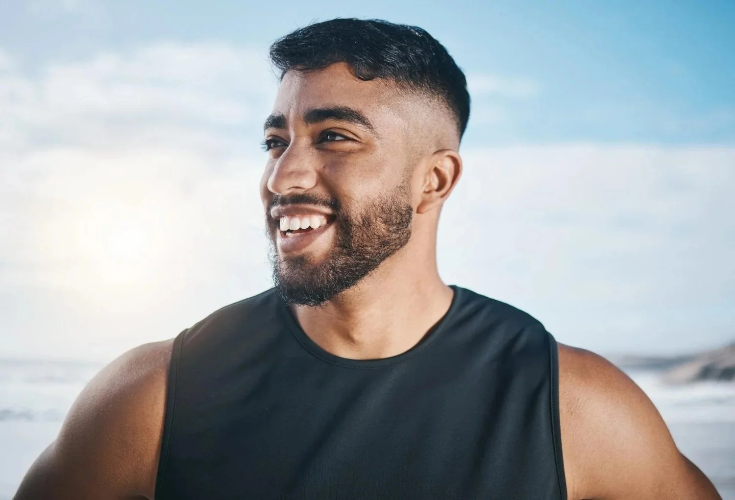 A smiling man with a short beard and a fade haircut at the beach, wearing a black sleeveless athletic shirt.