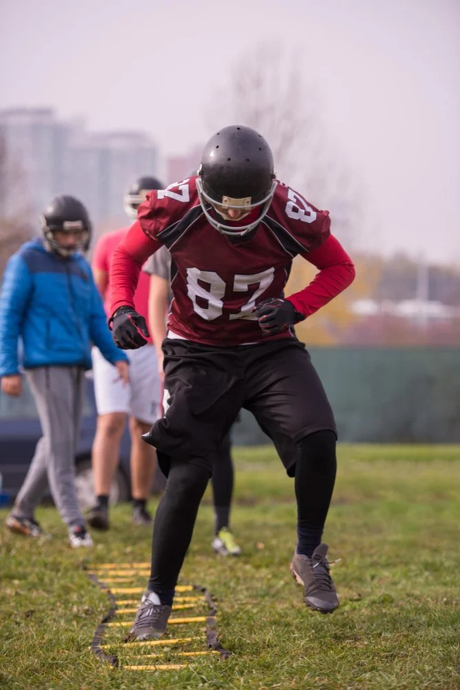 A football player in a red jersey with the number 87 practicing agility drills on a grassy field, with other players and coaches in the background.