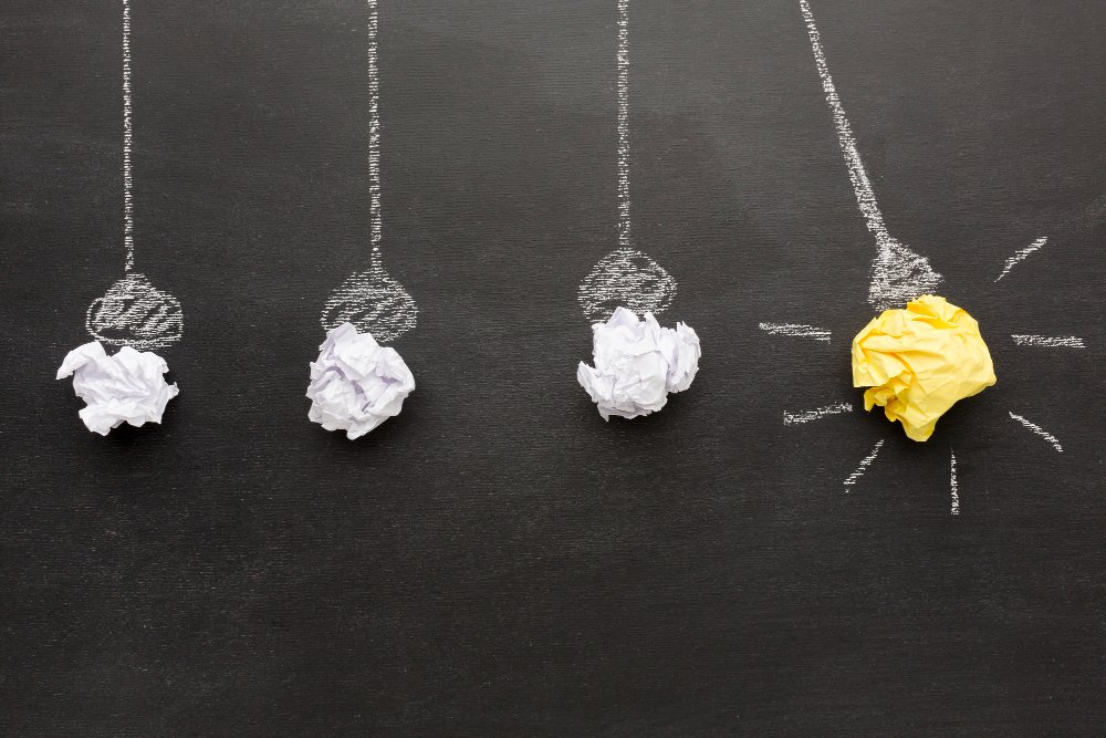 Chalk drawing of a Newton's cradle on a blackboard with four crumpled paper balls, one of which is yellow and represents a light bulb.
