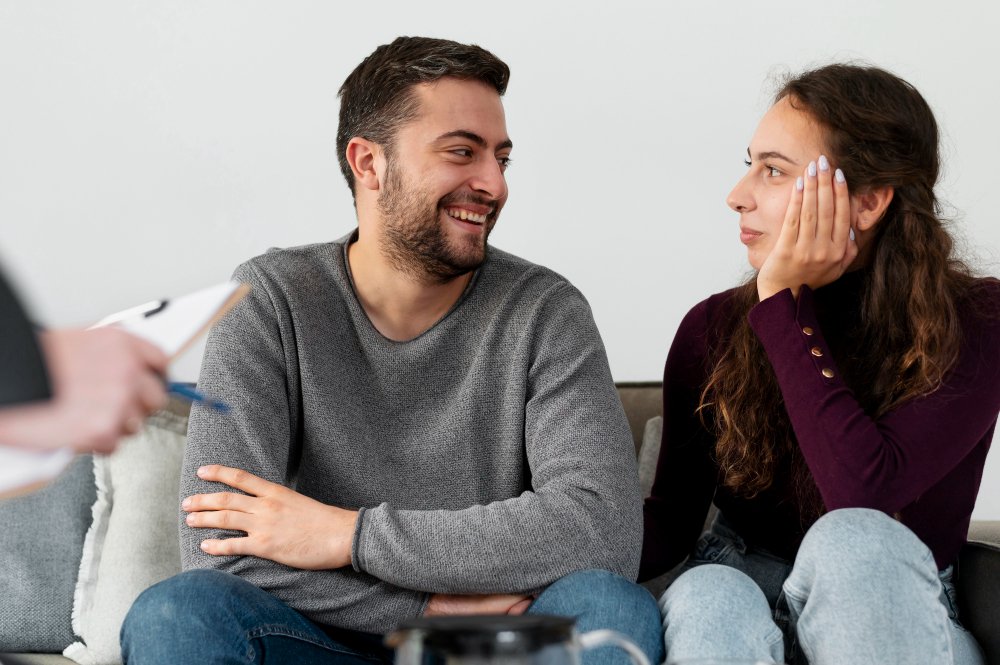 A man and woman sit on a couch during a therapy session or counseling appointment, engaging in conversation.