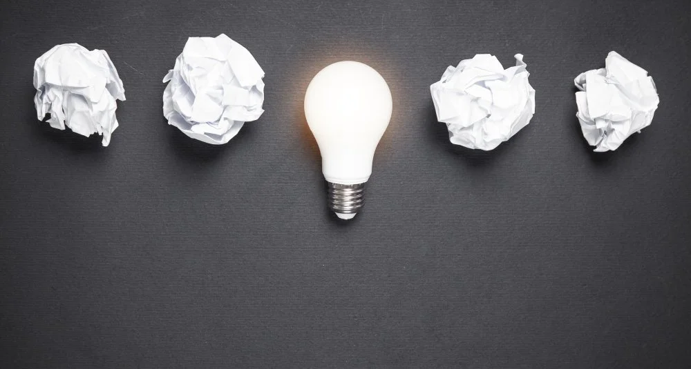 A row of five crumpled pieces of white paper surrounding a glowing light bulb on a dark surface.