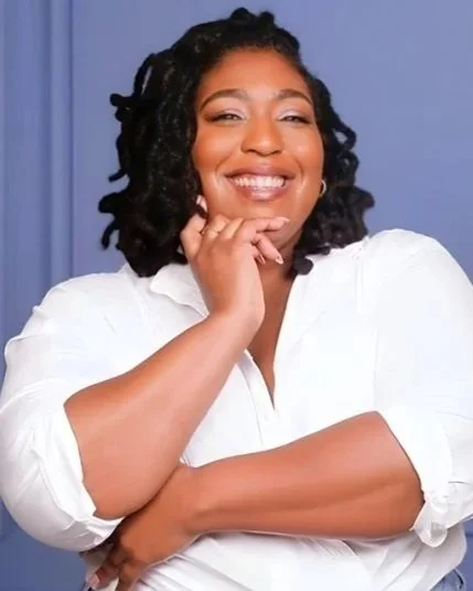 Woman with dark curly hair smiling, wearing a white blouse, with her hand near her chin, in front of a blue background.