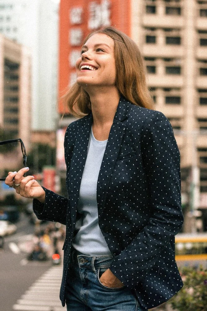 A woman with light brown hair smiling outdoors in an urban setting, holding glasses in her right hand, wearing a navy blazer with white polka dots and a white shirt.